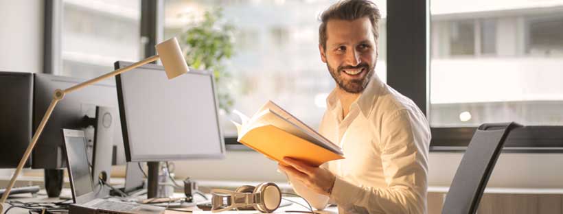 Homem sorrindo com livro na mão em frente ao computador- Home Office Produtivo - Impulso